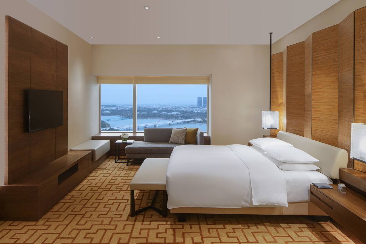 A modern hotel room with a large bed, a sofa, a TV, and a window with a city view.