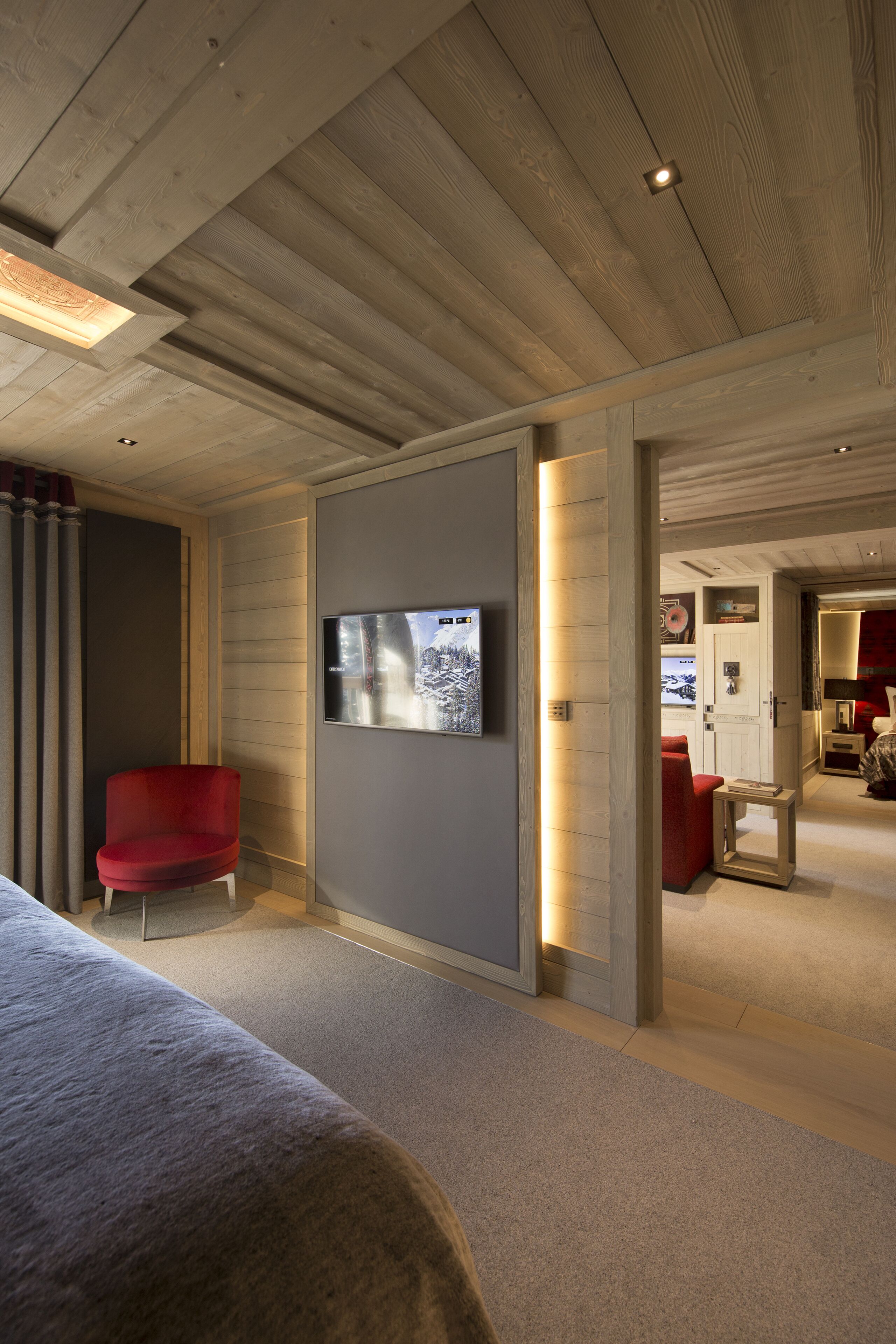A modern hotel room with a wooden ceiling, a flat-screen TV mounted on the wall, and a red armchair.