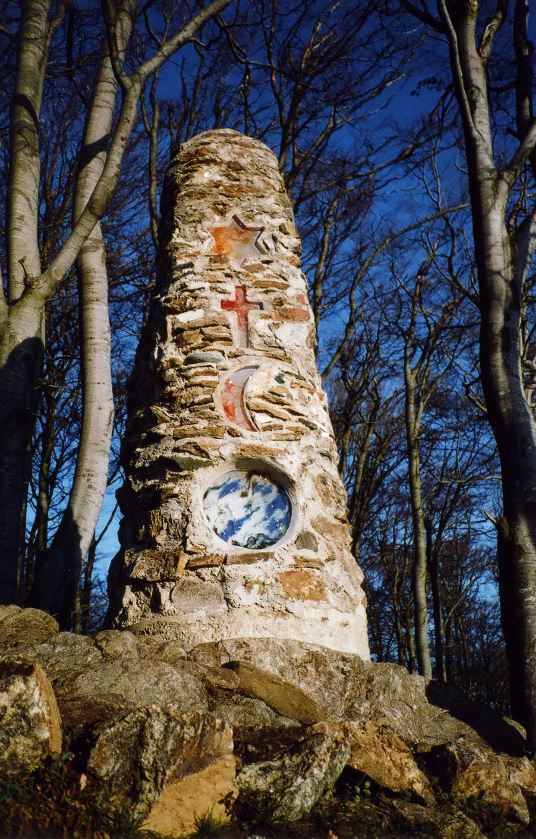 Monumento de piedra con relieves y una ventana circular, rodeado de árboles.