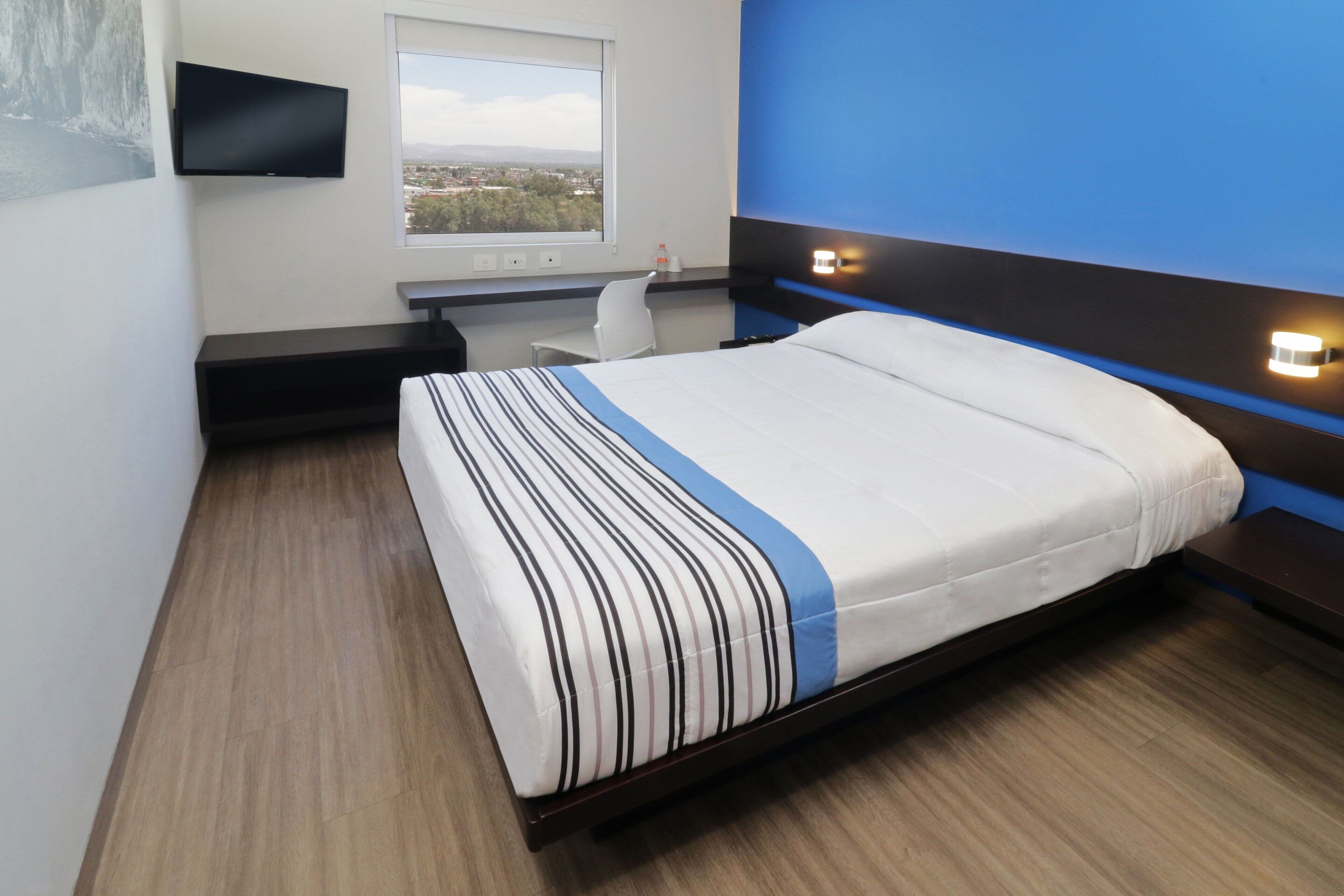 A modern hotel room with a bed, a desk, a chair, and a TV mounted on the wall.