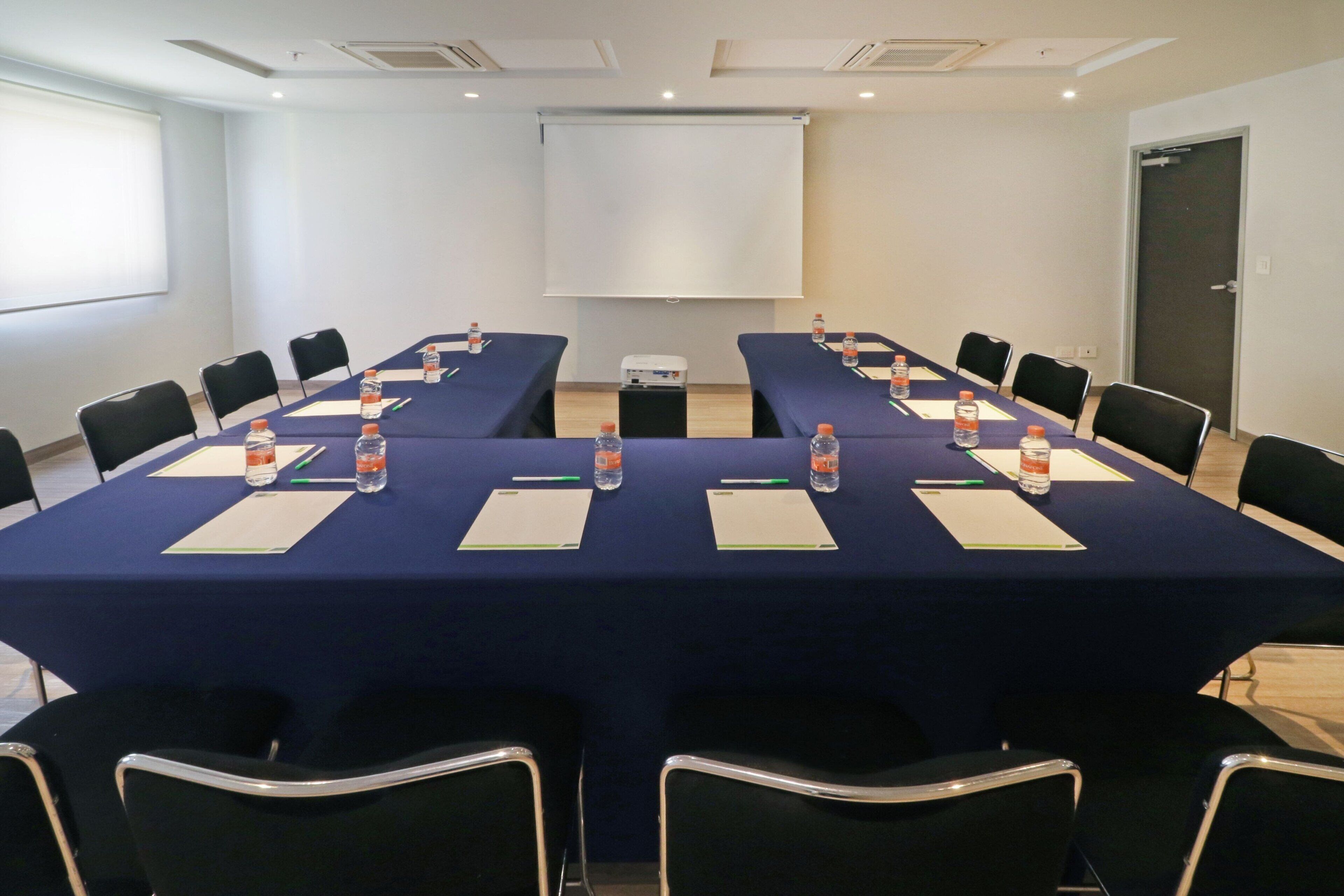 A conference room with a long table, chairs, water bottles, and a projector screen.