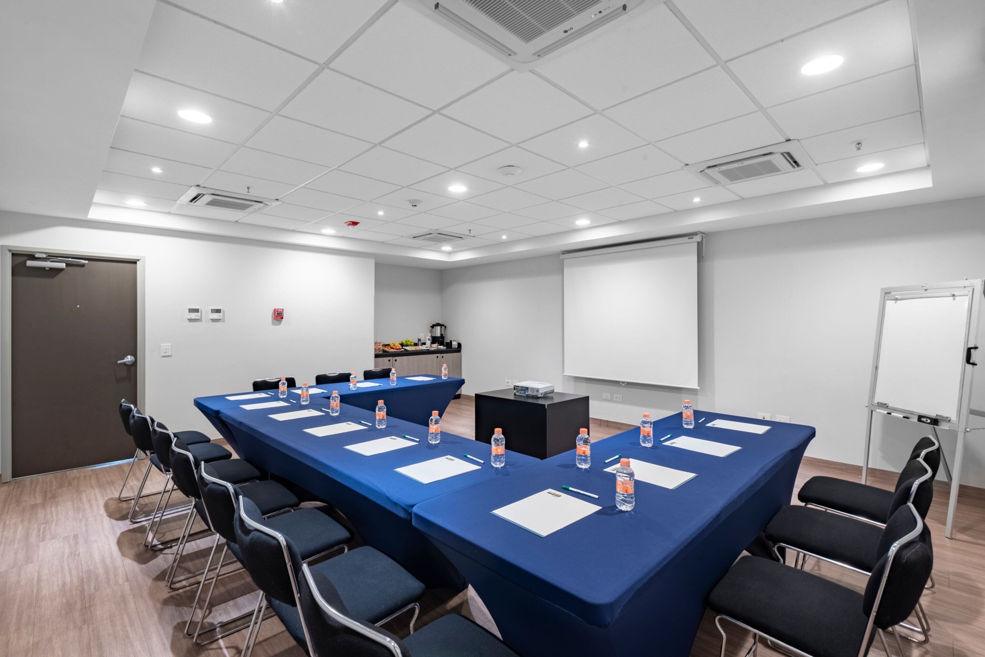 Un salón de conferencias con una larga mesa, sillas, una pantalla de proyector y una pizarra.