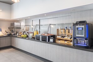 Un moderno mostrador de recepción de hotel con una máquina de café de autoservicio, una exhibición de frutas y una barra con diversos servicios.