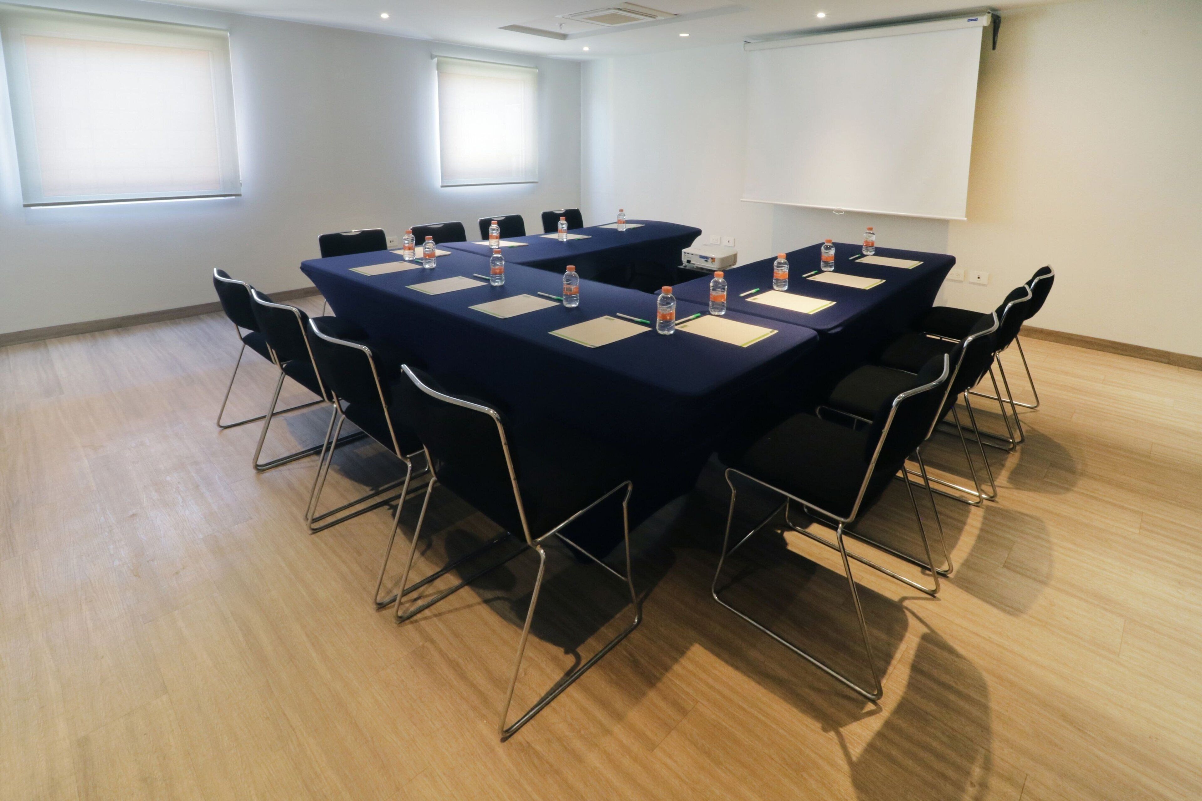 Una sala de conferencias con una gran mesa, sillas, botellas de agua y una pantalla de proyector.