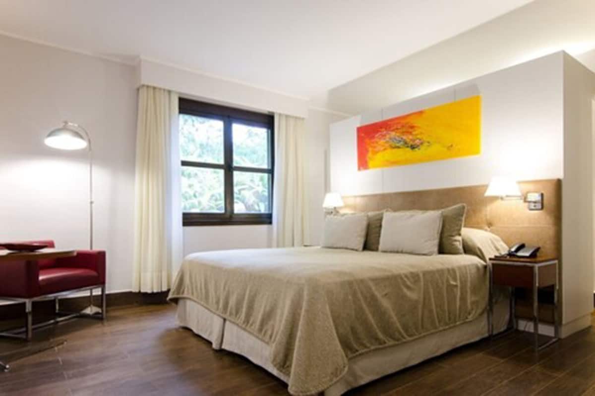 Ein Hotelzimmer mit Bett, Schreibtisch, Stuhl, Nachttisch, Lampe und Fenster.