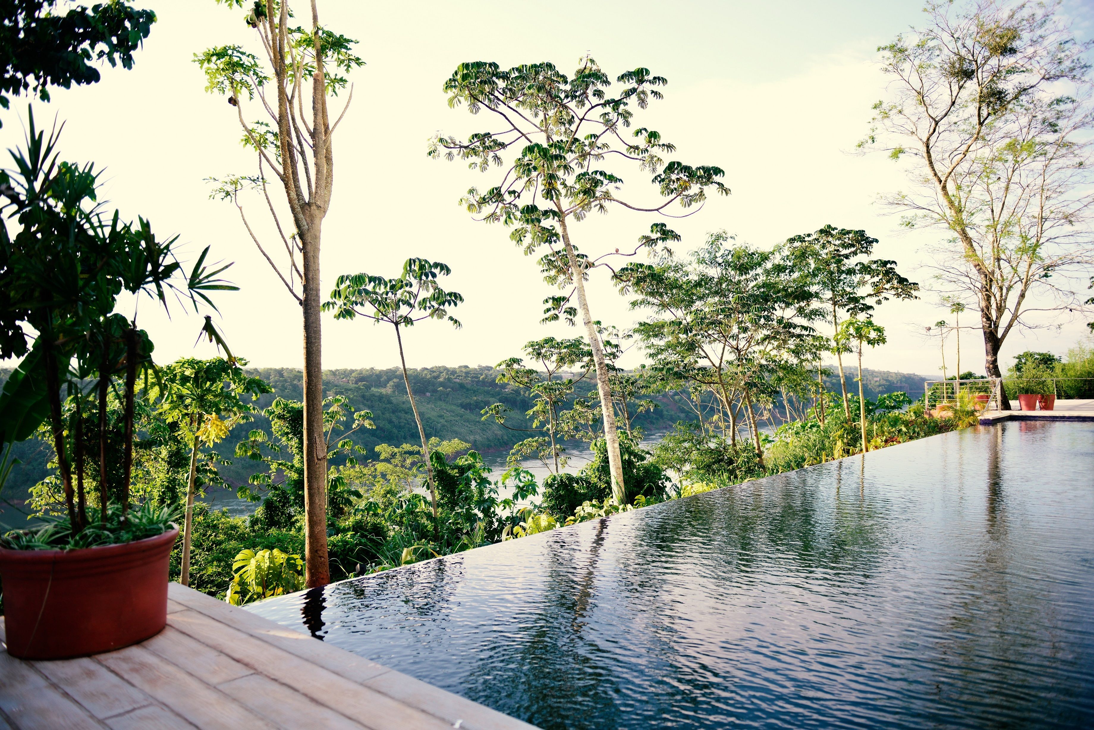 An infinity pool with a wooden deck, surrounded by lush greenery and trees, overlooking a hilly landscape.