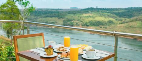 Una mesa con alimentos para el desayuno y un vaso de jugo de naranja, con vista a un río y colinas verdes.