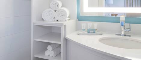 Un baño moderno con lavamanos blanco, espejo y repisas con toallas y artículos de aseo.