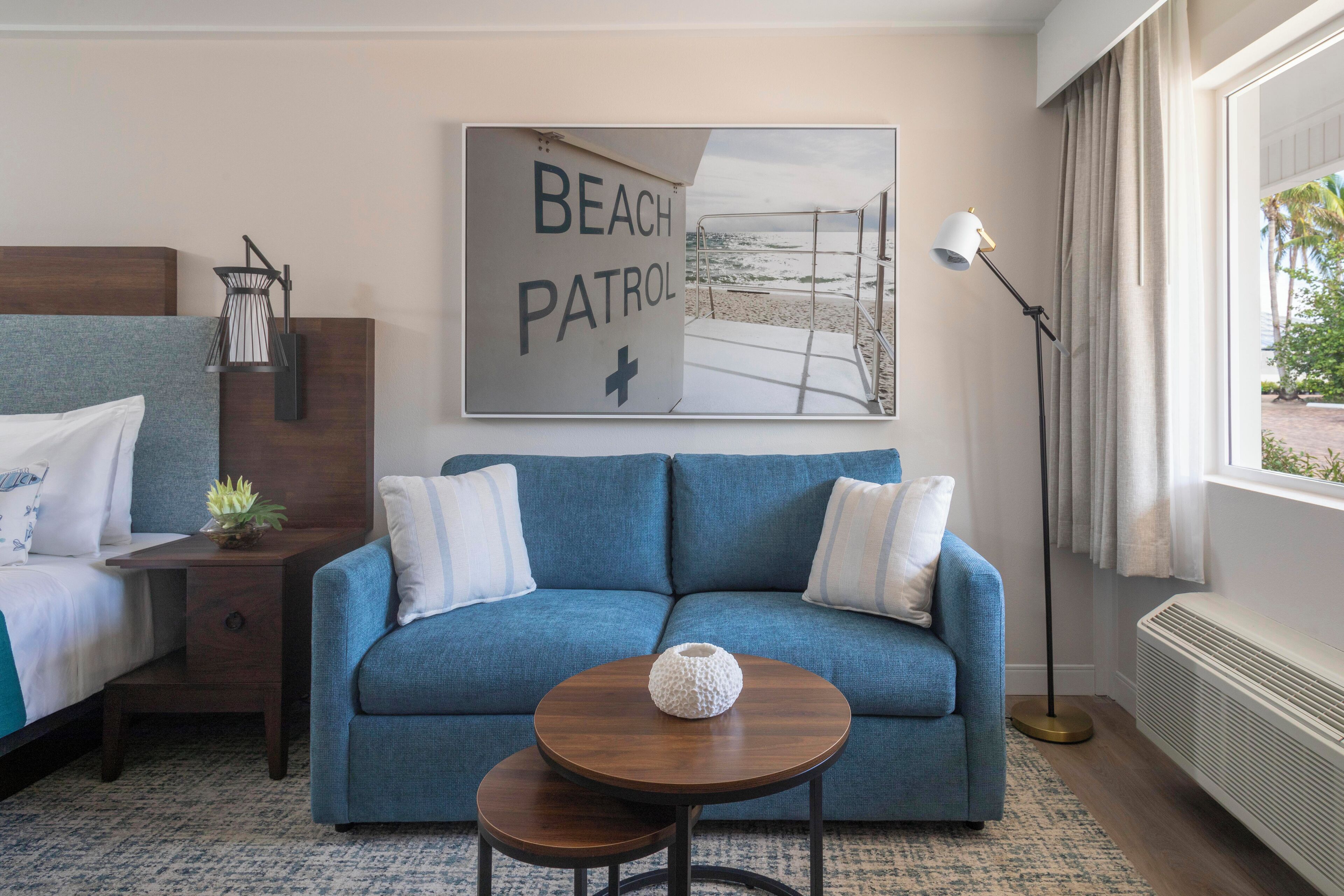A hotel room with a blue sofa, a small table with a vase, a bed with white bedding, a nightstand with a lamp, and a beach patrol sign on the wall.