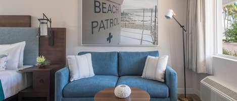 A hotel room with a blue sofa, a small table with a vase, a bed with white bedding, a nightstand with a lamp, and a beach patrol sign on the wall.