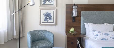 A hotel room with a bed, a chair, a round ottoman, a nightstand, a lamp, and two framed pictures on the wall.