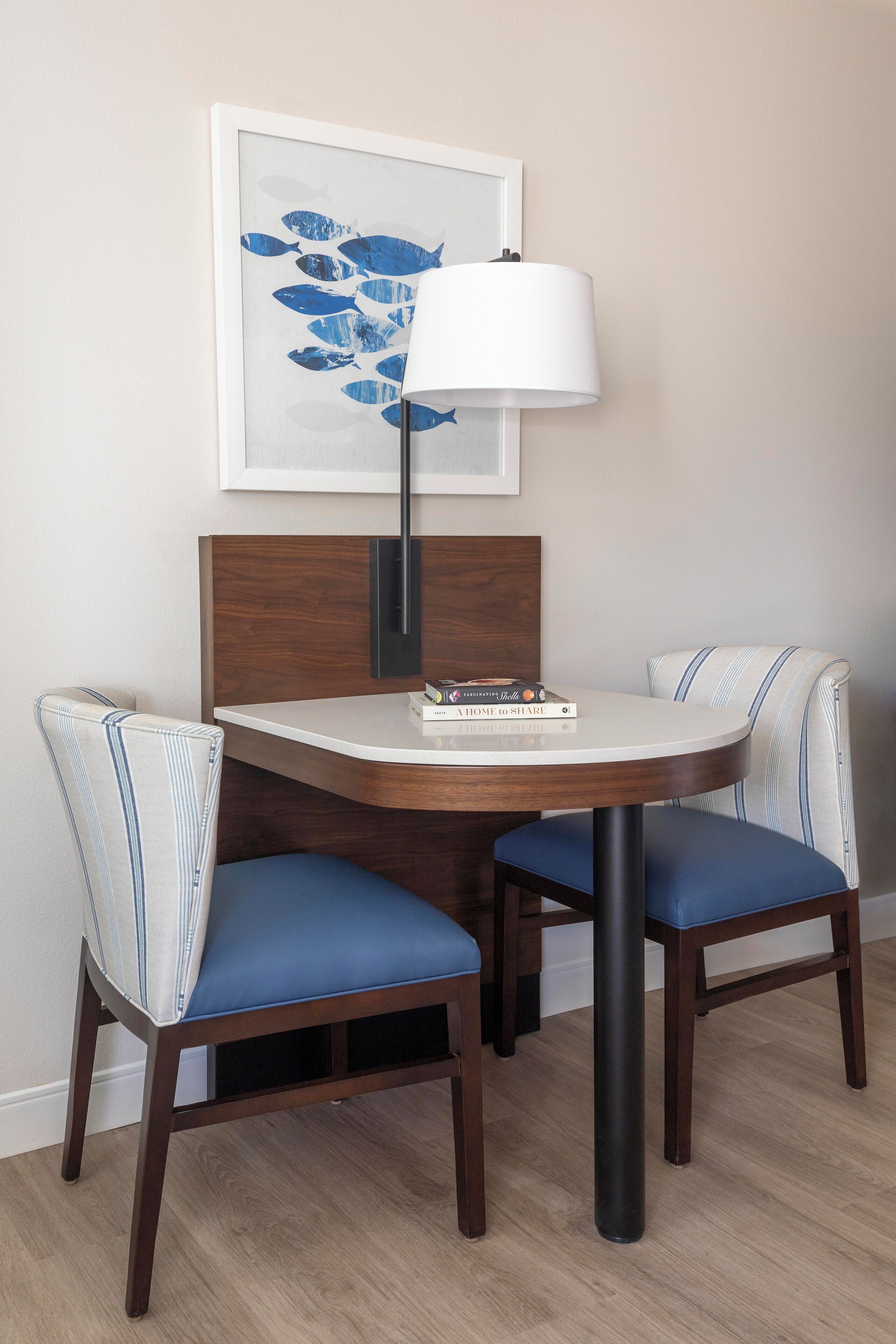 Een eetruimte met een ronde tafel, twee gestoffeerde stoelen met streepdessin, een wandlamp en een ingelijste kunstwerk erboven.