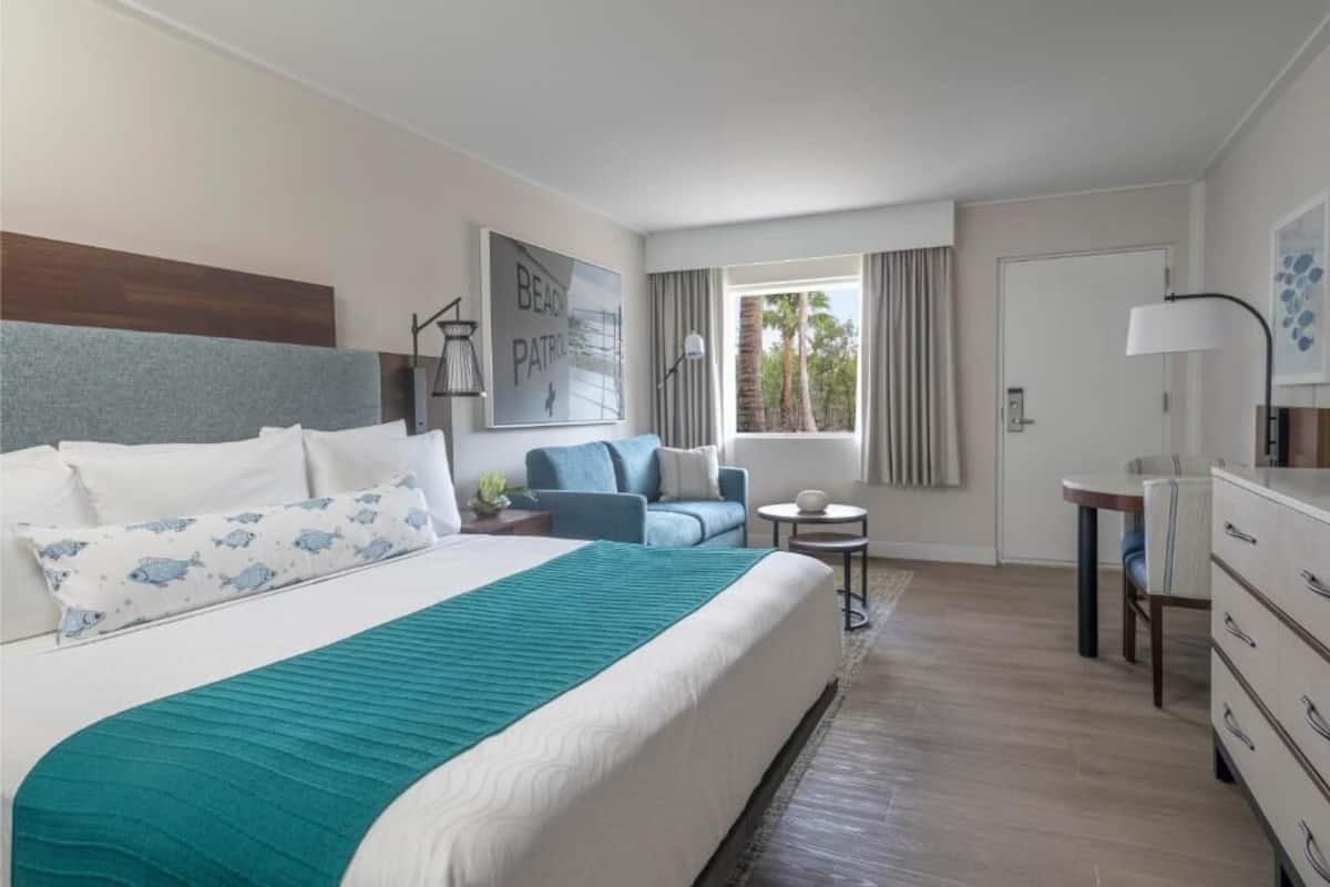 Habitación de hotel con una cama grande, una zona de estar con un sofá azul, una pequeña mesa y un escritorio con una lámpara.