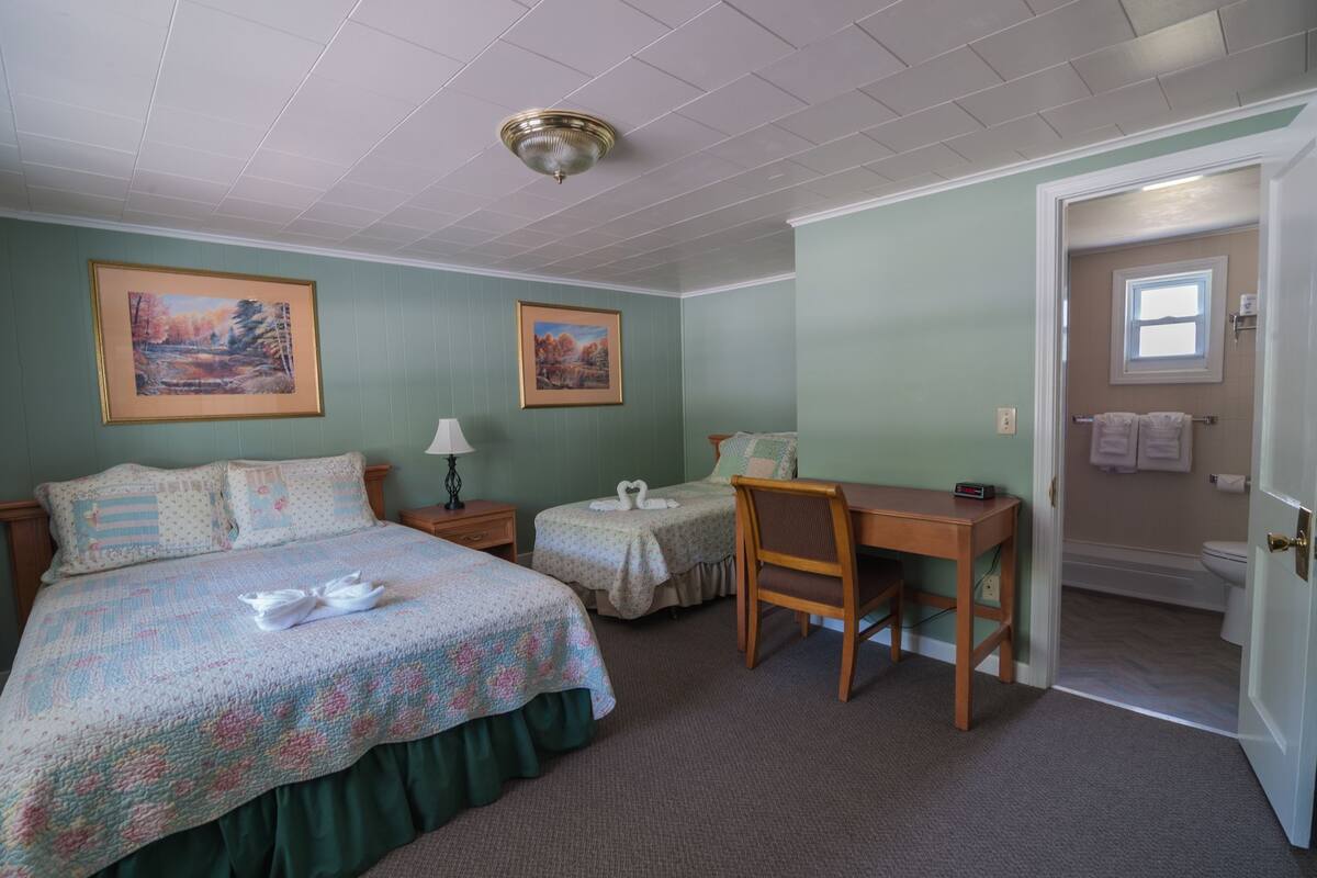 A bedroom with a bed, a desk, two chairs, a lamp, and two framed pictures on the wall.