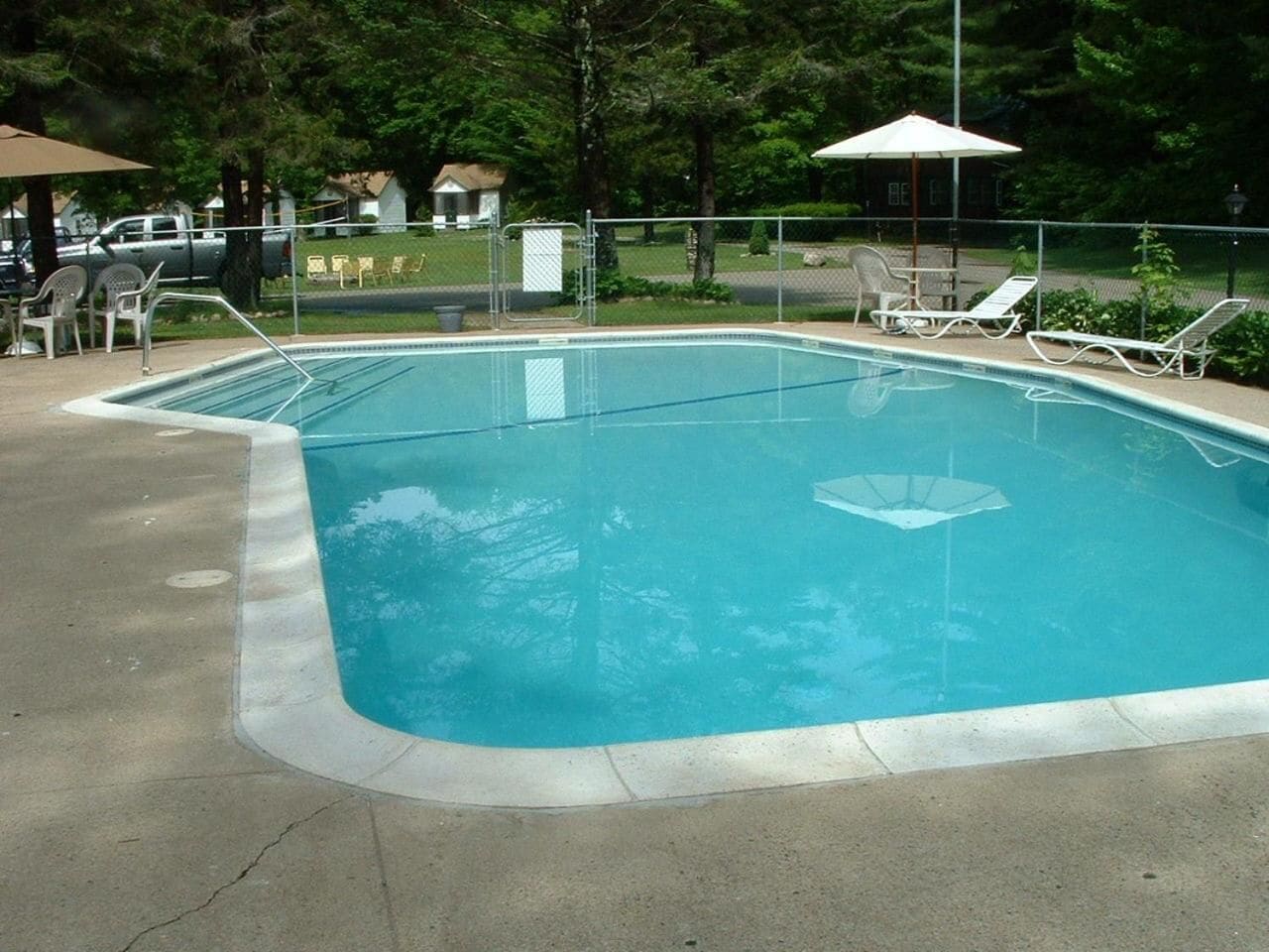 Piscina retangular com um deck de concreto, cercada por uma grade de metal. Há cadeiras de espreguiçadeira e guarda-sóis ao longo da borda da piscina.