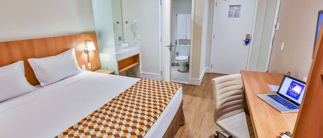 Quarto de hotel com uma cama, uma escrivaninha com um laptop, uma cadeira, um banheiro e uma porta.