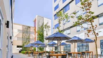 Uma área de café ao ar livre com mesas e cadeiras, guarda-sóis azuis e um edifício com vários andares e janelas ao fundo.