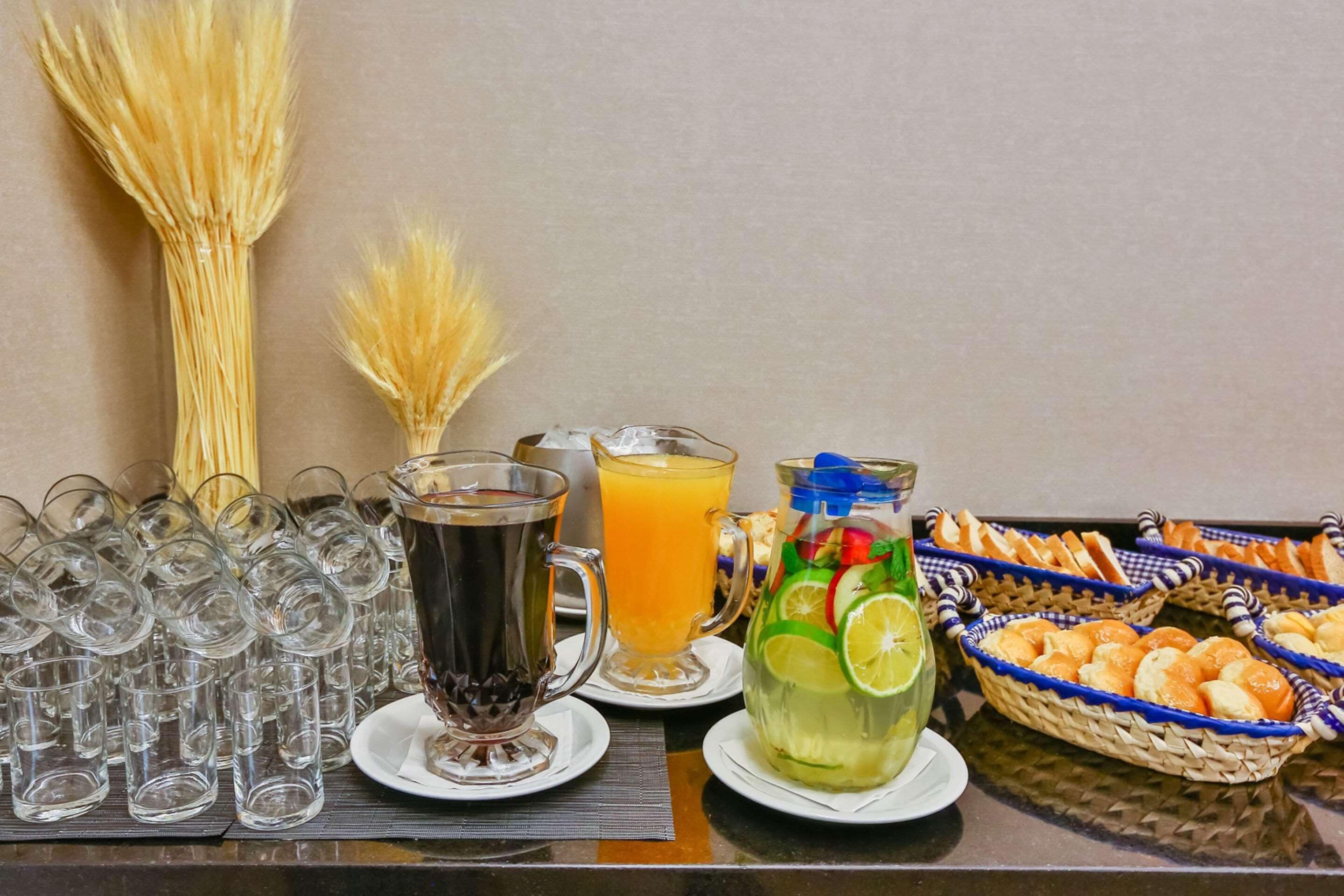 A buffet table with various beverages, including a pitcher of iced tea, a glass of orange juice, and a pitcher of iced tea with lime slices. There are also baskets of bread and a large bunch of wheat stalks in the background.