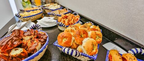 A buffet table with various food items including bread, pastries, and a drink.