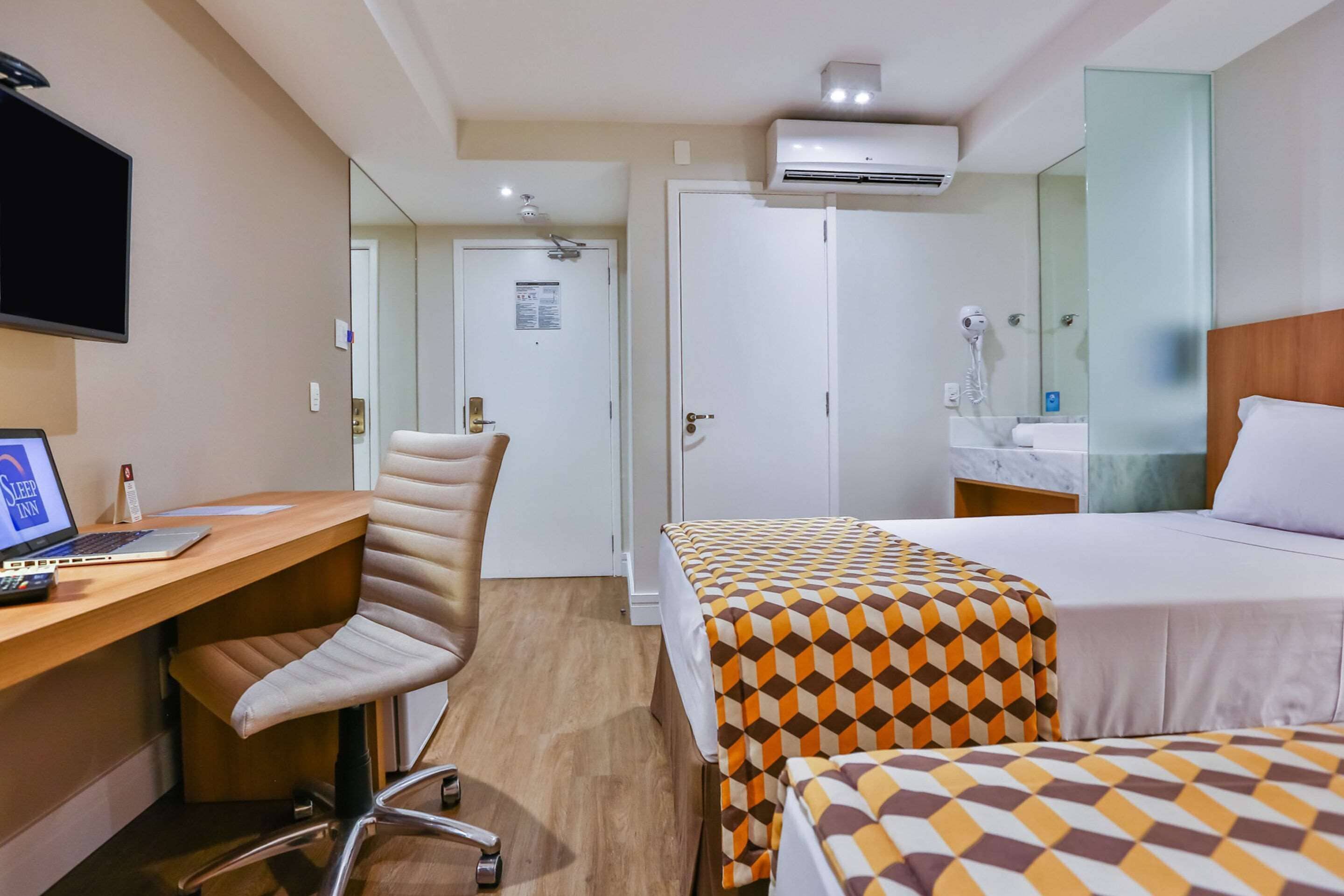 Habitación de hotel con escritorio, silla, dos camas, televisor, aire acondicionado, espejo y baño con lavamanos y secador de pelo.
