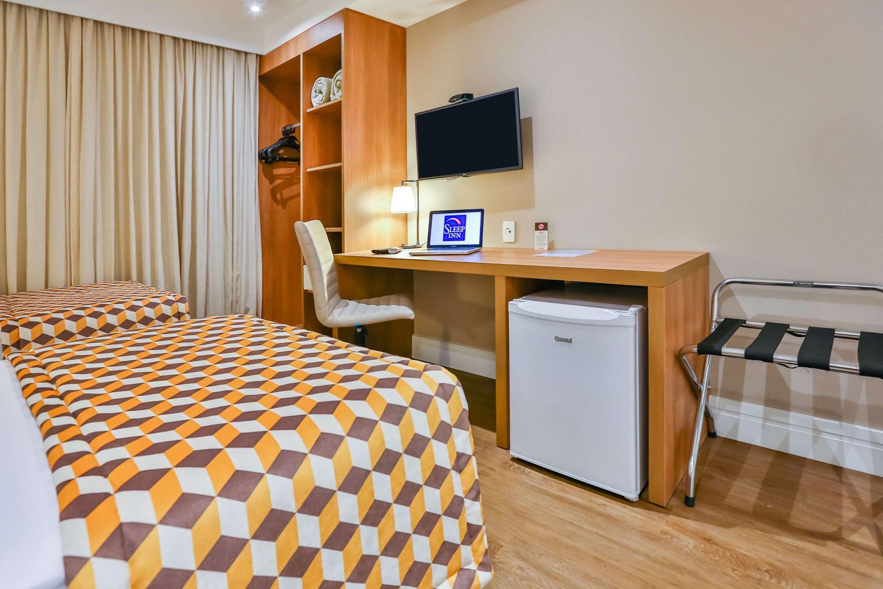 A hotel room with two beds, a desk with a computer, a television, and a refrigerator.