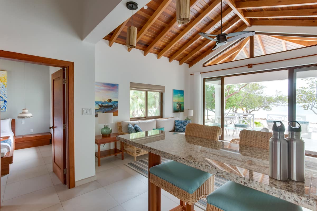 A spacious living area with a high ceiling, wooden beams, a large dining table, wicker furniture, and large windows offering a view of the outdoors.