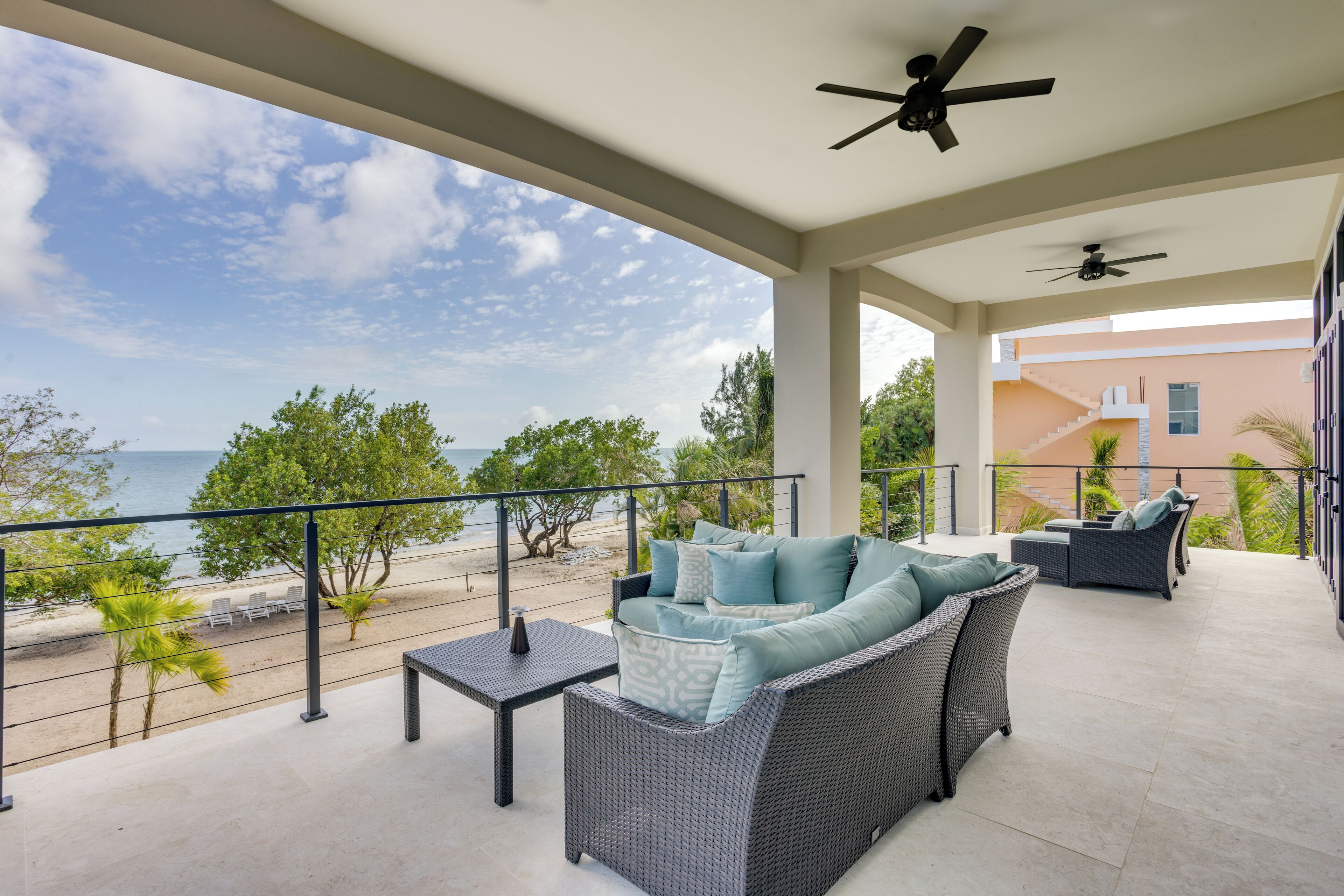 Un espace extérieur aménagé, abrité et doté de sièges, offrant une vue sur la plage et l’océan, avec des meubles en osier et des ventilateurs de plafond.