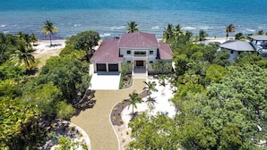 A two-story house with a red roof, surrounded by lush greenery and a beach.