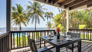 A wooden deck with a table and chairs overlooking a beach with palm trees and a view of the ocean.