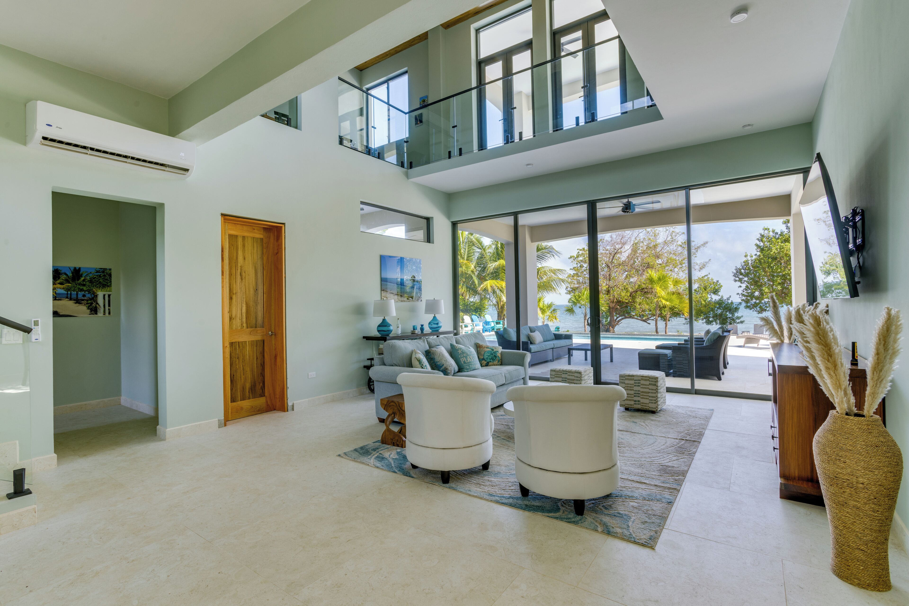 A spacious living area with a high ceiling, a glass balcony, a large window offering a view of palm trees, and a modern sofa set.