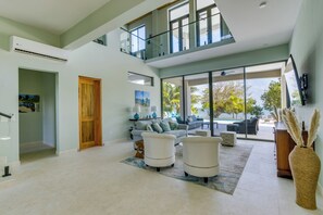 A spacious living area with a high ceiling, a glass balcony, a large window offering a view of palm trees, and a modern sofa set.