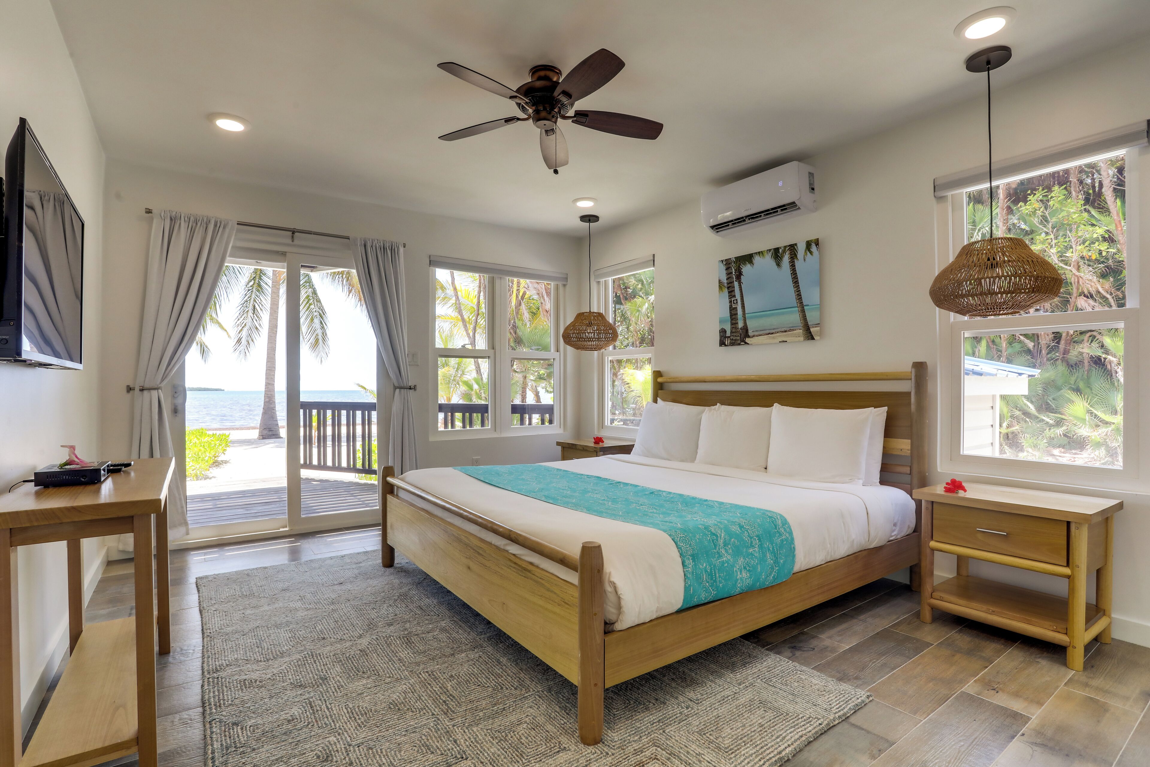 A bedroom with a wooden bed, a ceiling fan, a nightstand, a desk, and a large window with a view of palm trees.
