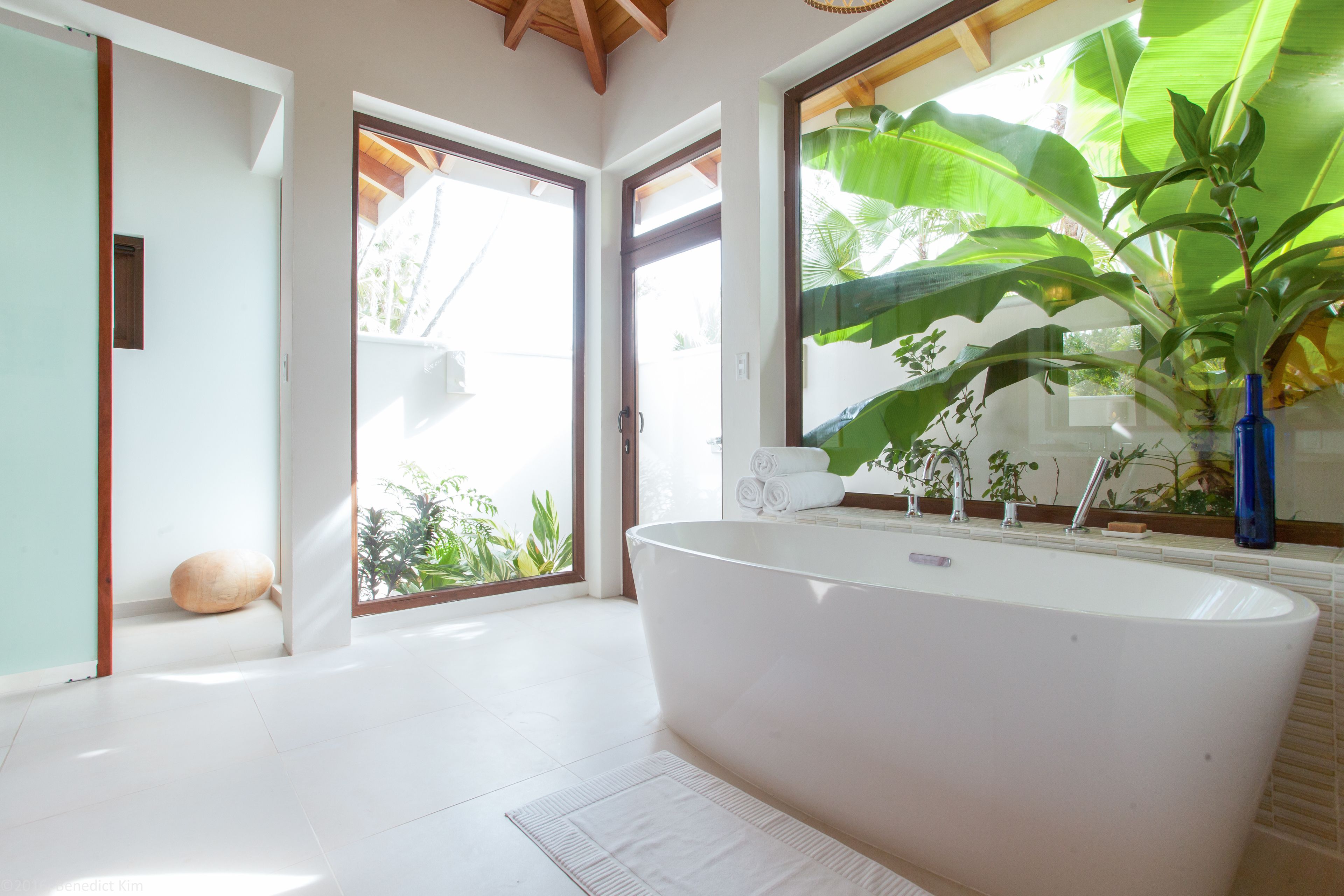 A modern bathroom with a freestanding bathtub, a large window with a view of greenery, and a wooden floor.