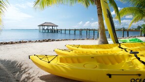 A beach with yellow kayaks, a palm tree, and a thatched-roof structure in the distance.
