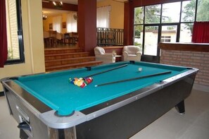 A pool table with balls and cues, a staircase leading to a dining area, and a view of the outside through large windows. - Hotel Taormina (Villa Carlos Paz)