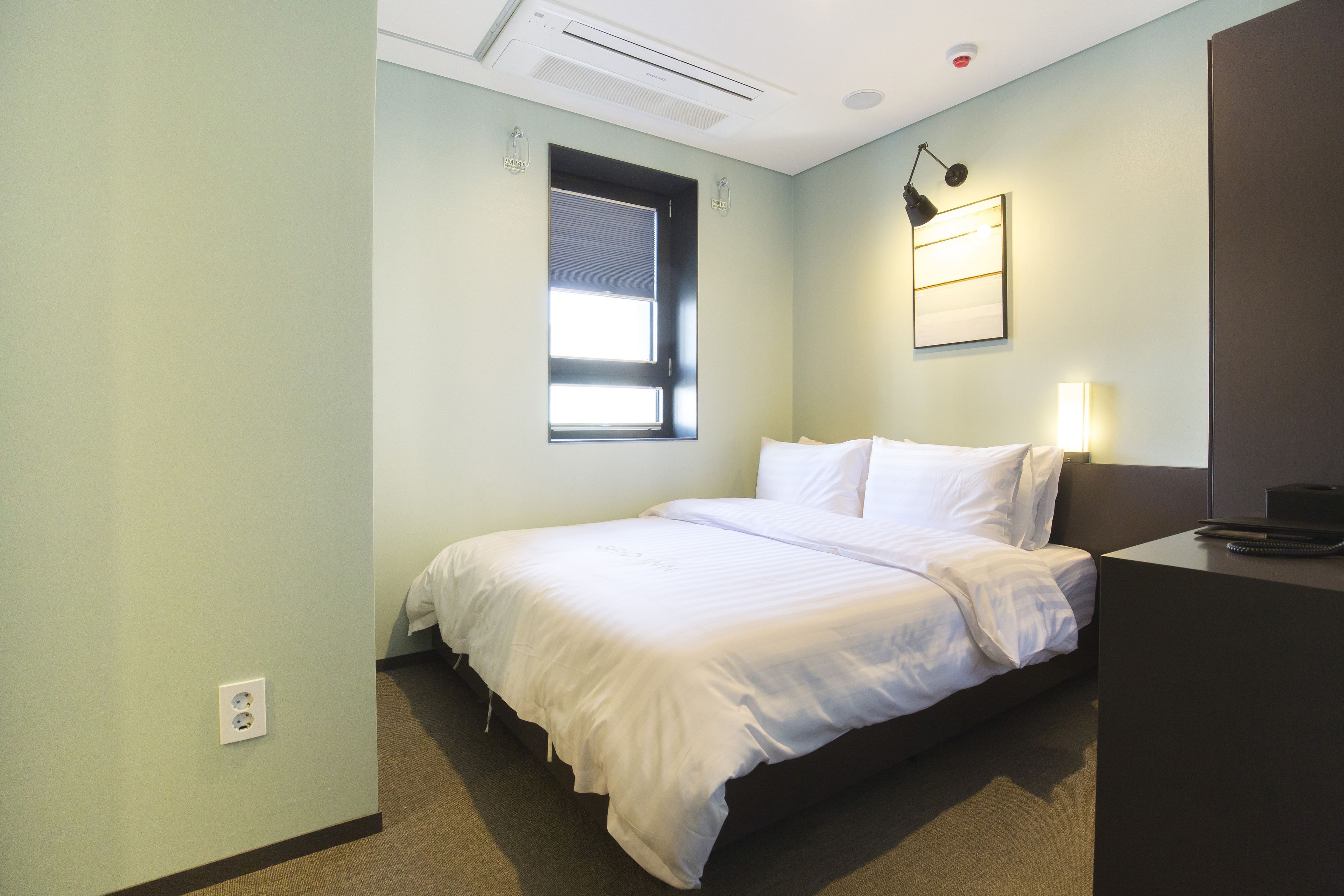A neatly made bed with white linens, a bedside table with a lamp, and a window with blinds.