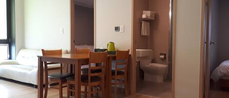 A modern living room with a wooden dining table and chairs, a sofa, and a view of a bathroom with a toilet and shower.