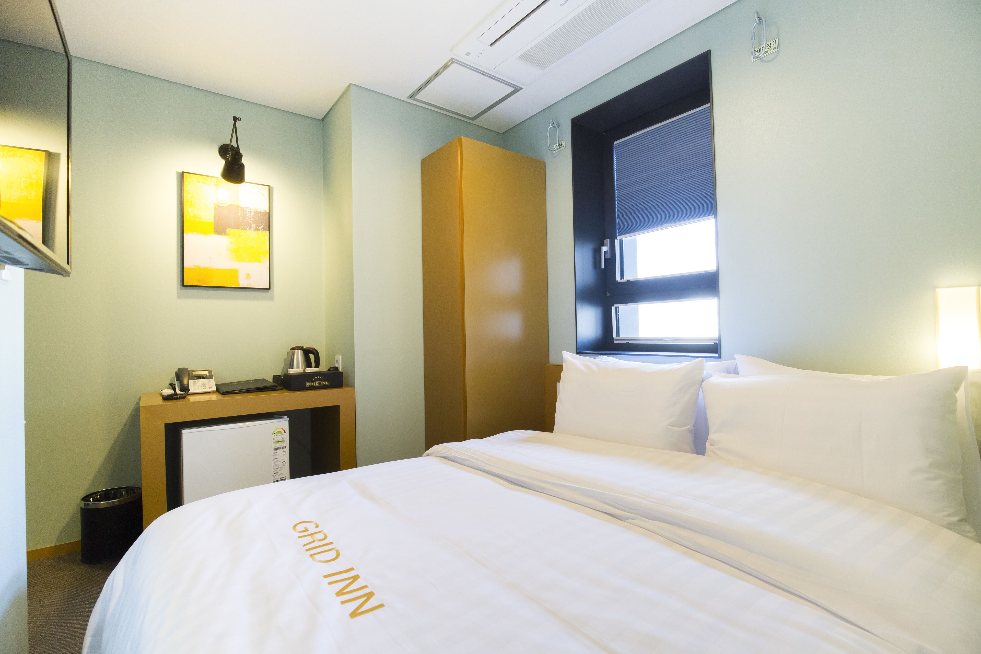 A hotel room with a bed, a desk with a phone and a coffee maker, a small trash bin, and a window with blinds.