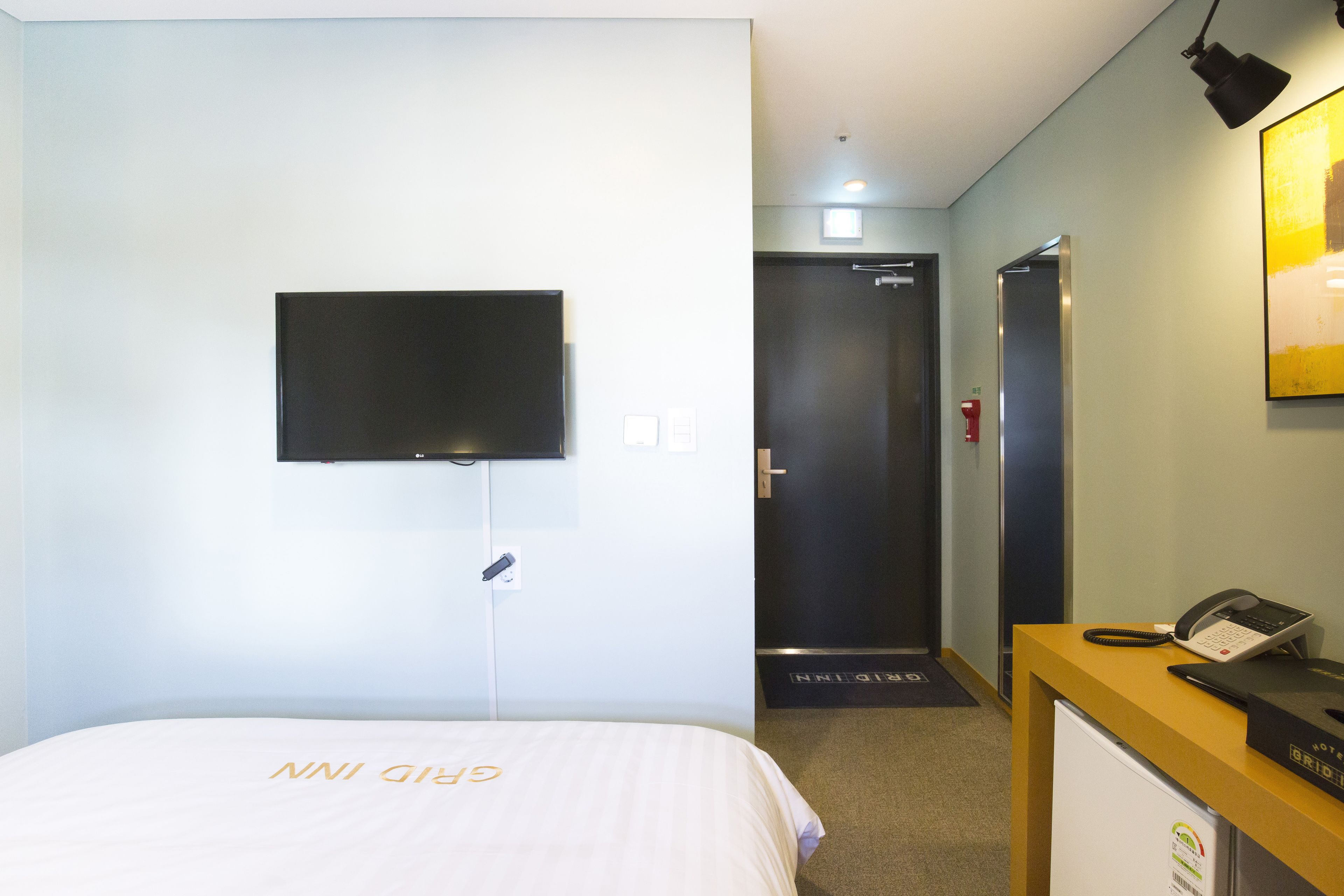 Ein Hotelzimmer mit Bett, einem an der Wand befestigten Fernseher, einem Schreibtisch mit Telefon und einer Tür zu einem Badezimmer.