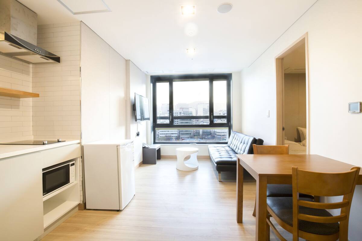 A modern kitchen with white appliances, a dining area with a wooden table and chairs, and a large window offering a city view.
