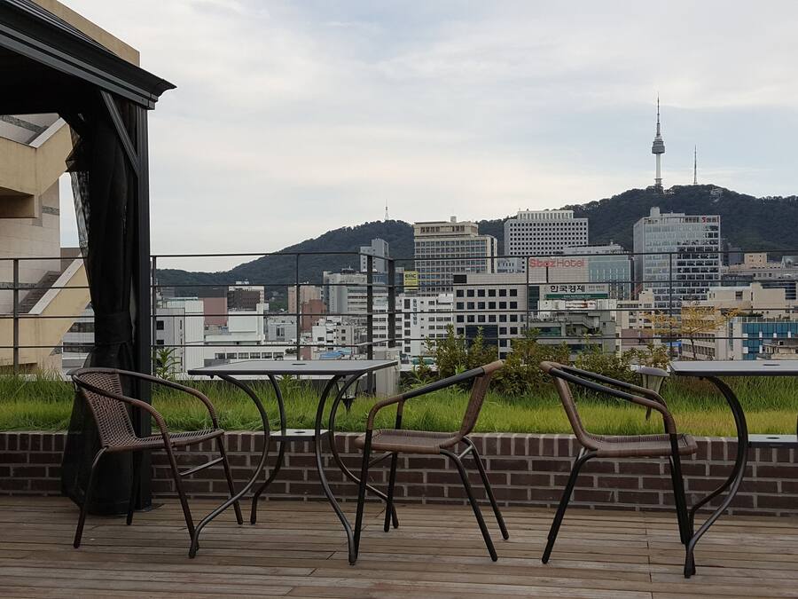 Terraza al aire libre con sillas y mesas de mimbre, con vistas a una ciudad que muestra edificios modernos y una torre destacada.