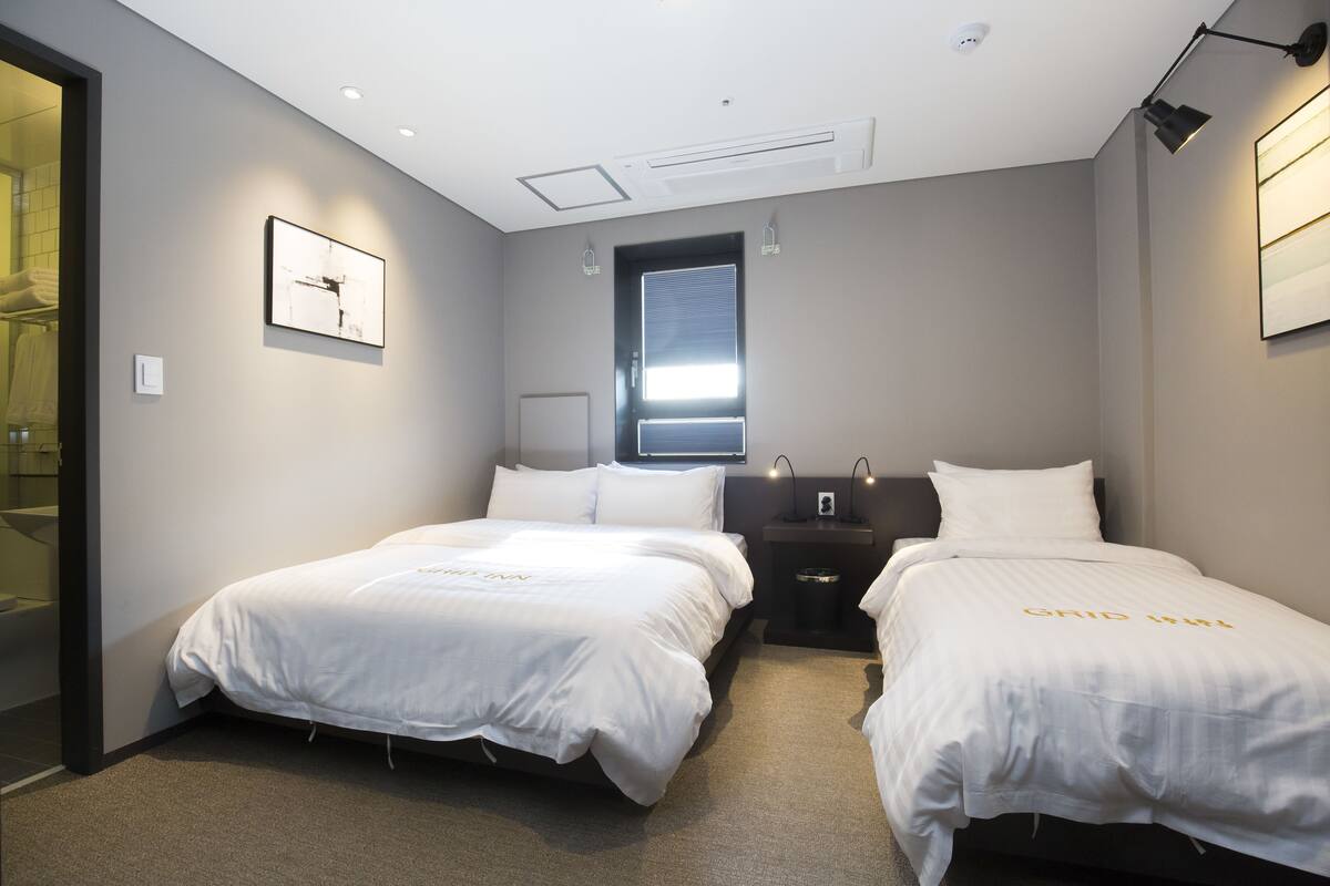 A hotel room with two beds, a window, a bedside table, and a wall-mounted light fixture.