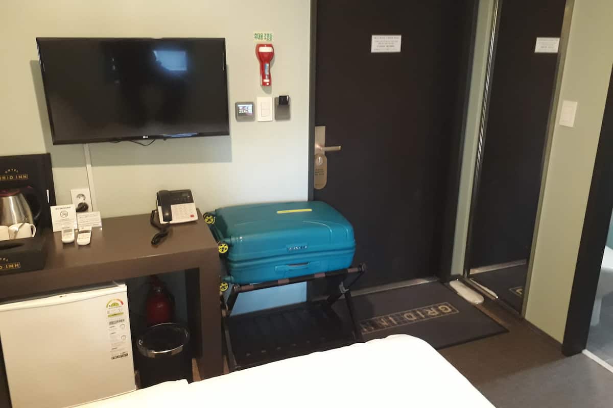 A hotel room with a bed, a desk with a television, a minibar, a suitcase, and a door with a handle.