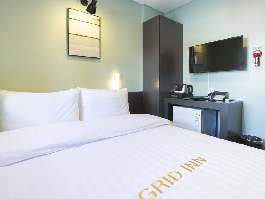 A hotel room with a bed, a desk with a telephone, a television, and a wall-mounted light fixture.