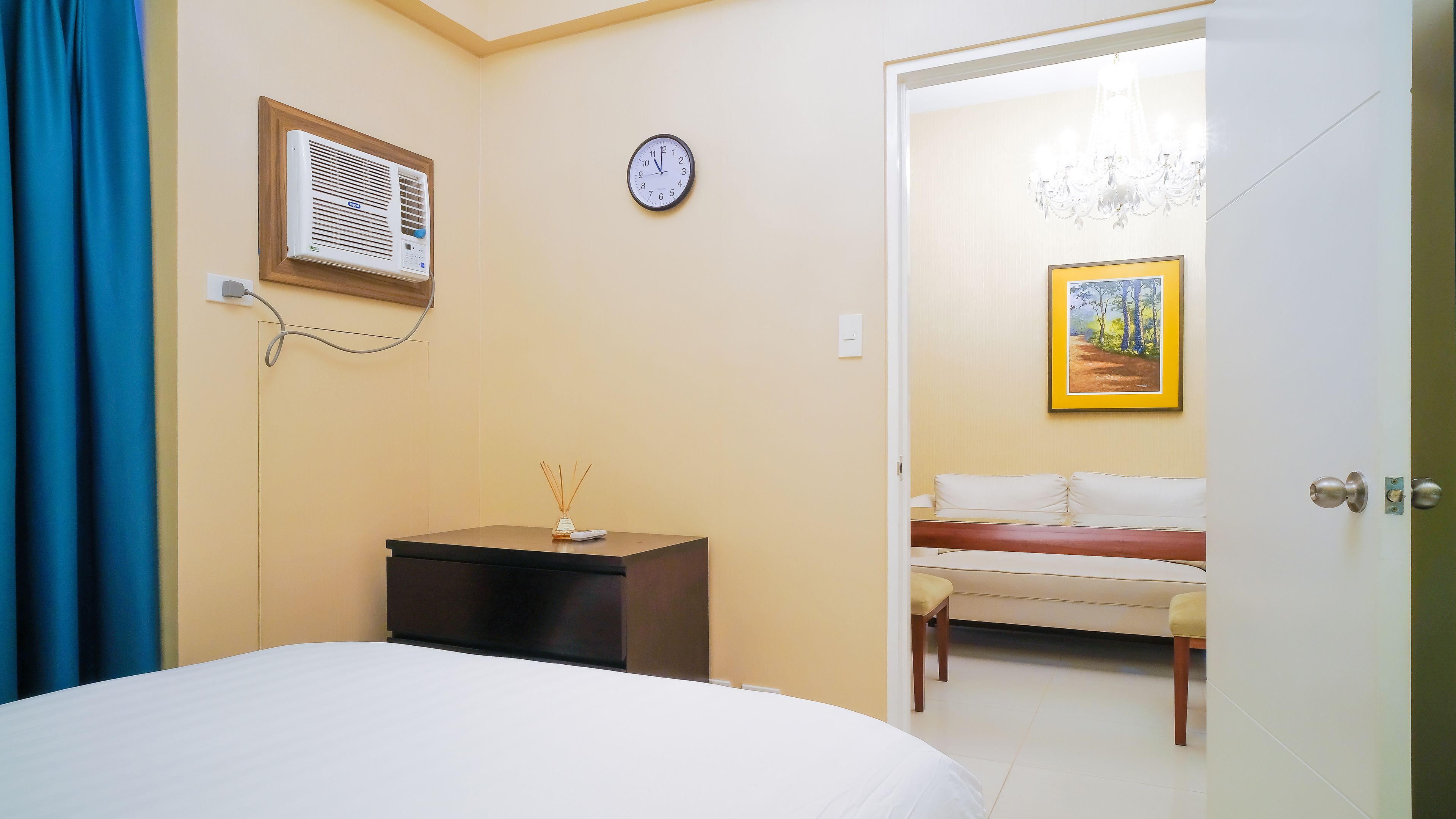 A bedroom with a bed, a bedside table with a lamp, a wall clock, and a door leading to another room.