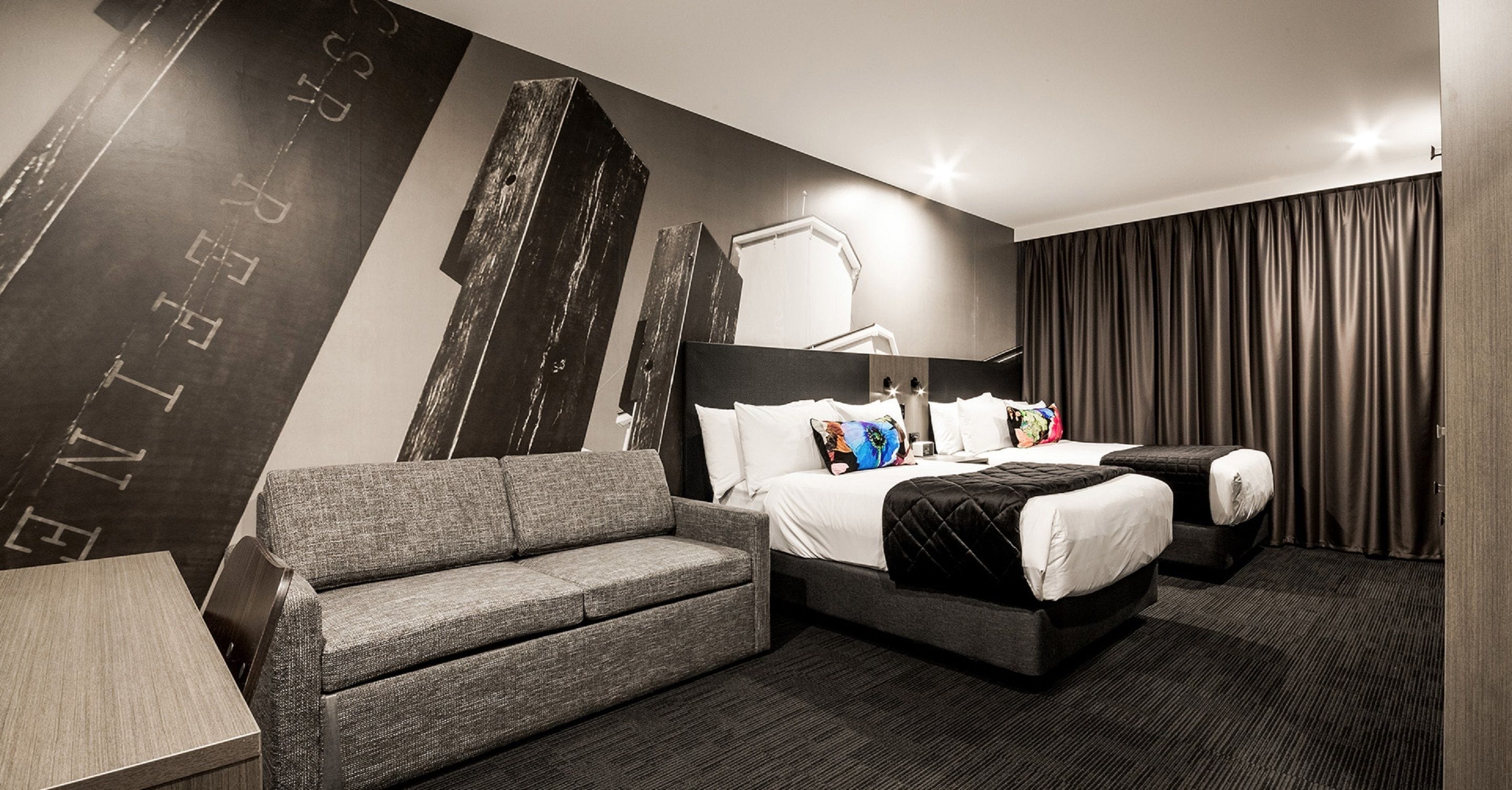 A modern hotel room with a large bed, a grey sofa, a desk, and a dark patterned wall.