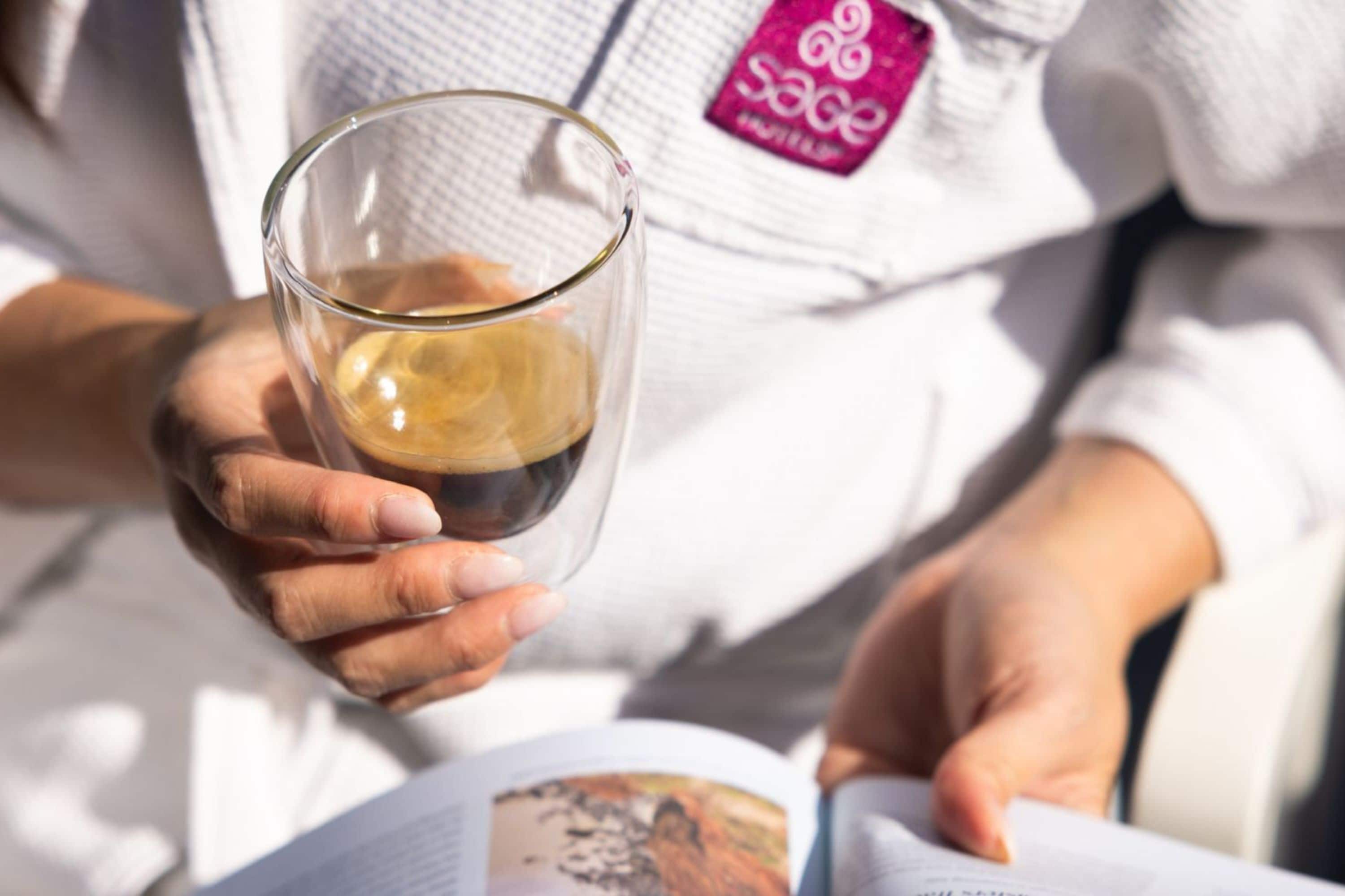 Iemand houdt een koffiekop vast, draagt een witte trui met het logo 'Sage' en leest een tijdschrift.