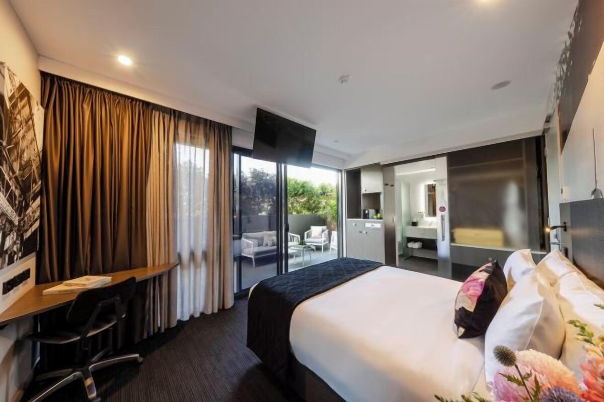 Habitación de hotel con cama, escritorio, silla, televisor y puerta corrediza a un balcón.