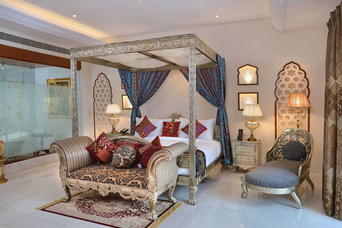 A four-poster canopy bed with ornate bedding, a chaise lounge, and a nightstand with lamps.