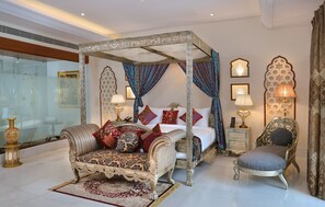 A four-poster canopy bed with ornate bedding, a chaise lounge, and a nightstand with lamps. - Indana Palace Jaipur (Amer)
